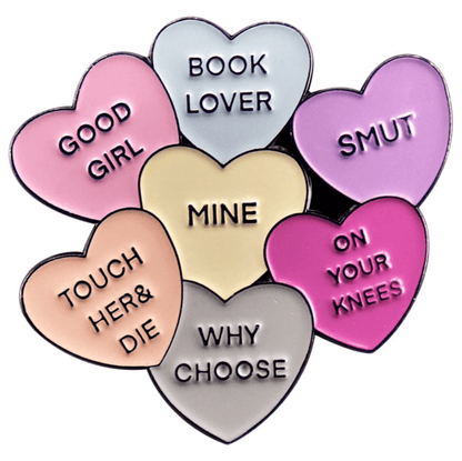 A cluster of seven pastel love hearts enamel pins, each with unique text: GOOD GIRL, SMUT pin, BOOK LOVER, ON YOUR KNEES, WHY CHOOSE, TOUCH HER & DIE, and MINE at the center.