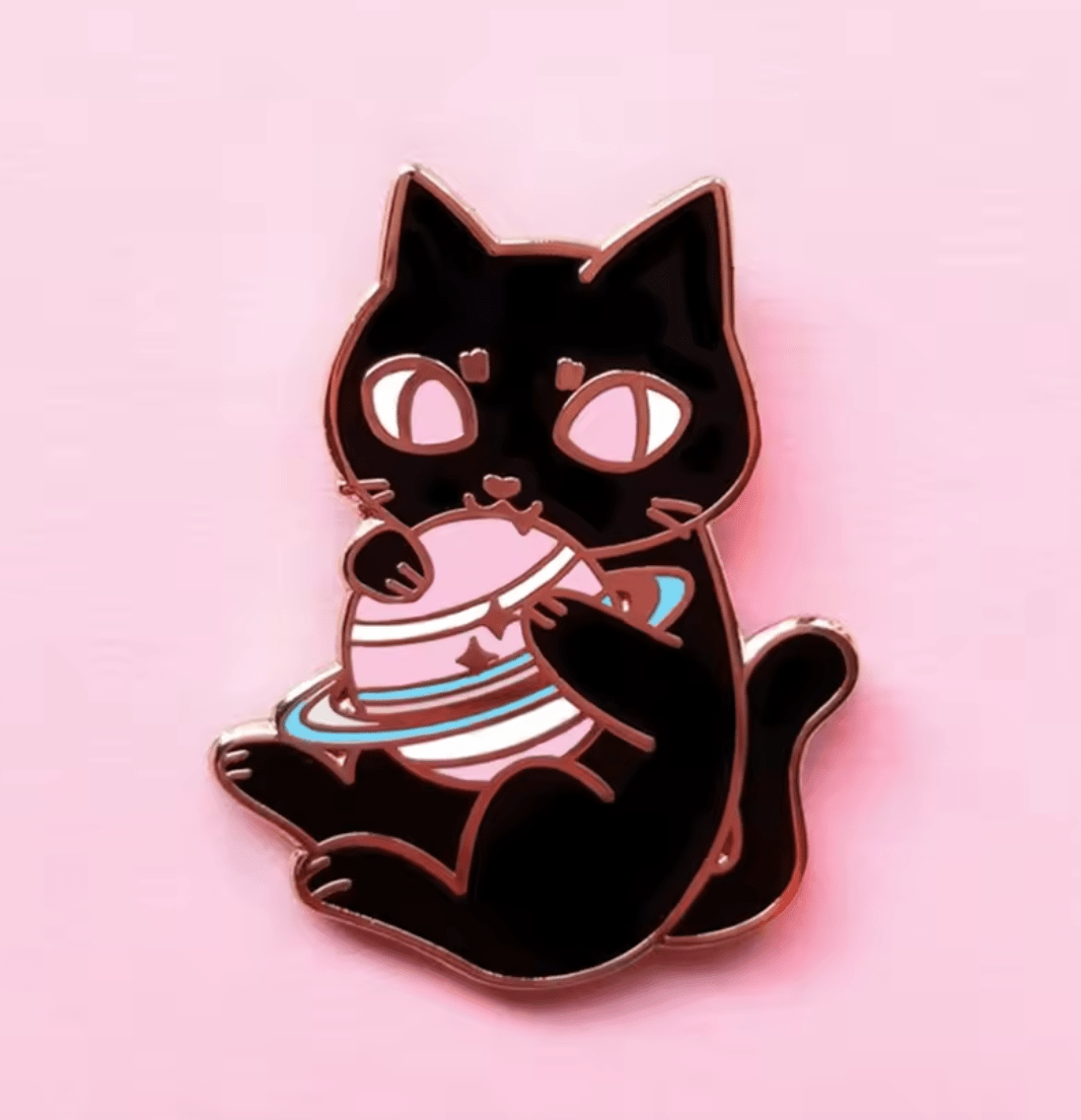 black cat enamel pin shows a wide-eyed cat on a pink background, playfully holding a Saturn-like planet with rings—a whimsical, cosmic accessory for space lovers.
