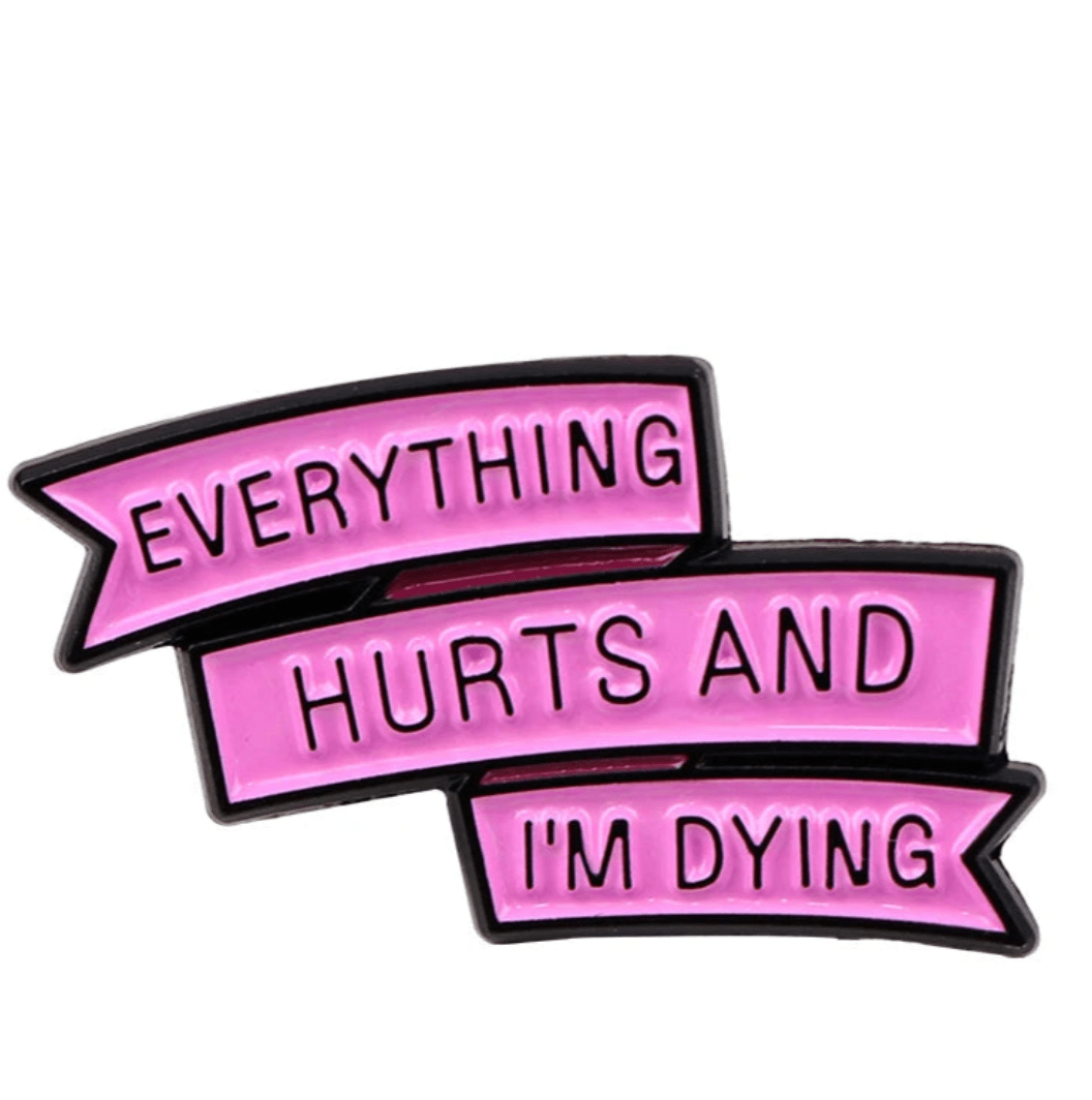 The Kiss Me Later enamel pin features a pink ribbon with three sections reading “EVERYTHING HURTS AND IM DYING” in bold black letters. It has a black outline and a playful, comic style.