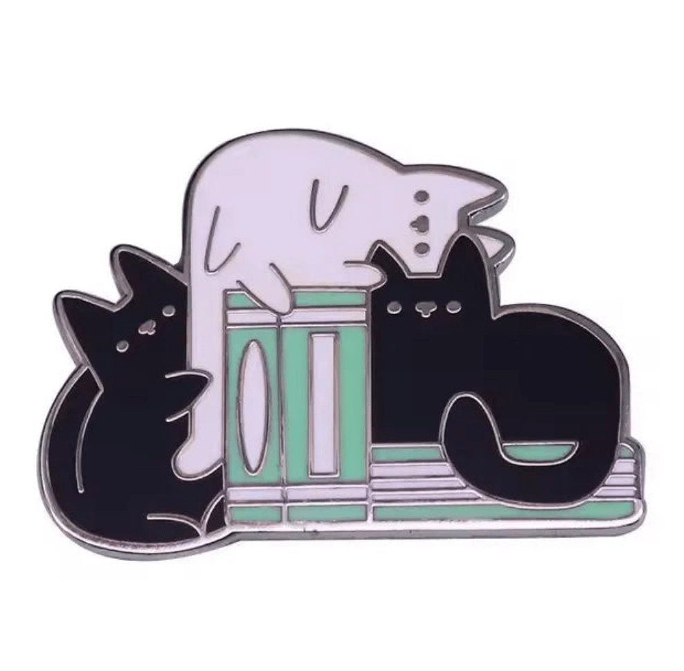enamel pin features two black cats and one white cat lounging on mint green and pink books—a charming gift for book lovers or collectors of book-themed pins.