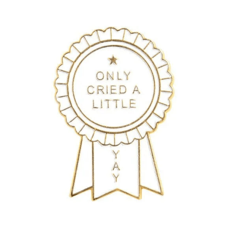 A gold and white enamel pin, shaped like an award ribbon with ONLY CRIED A LITTLE in the center and YAY on the hanging ribbons below.