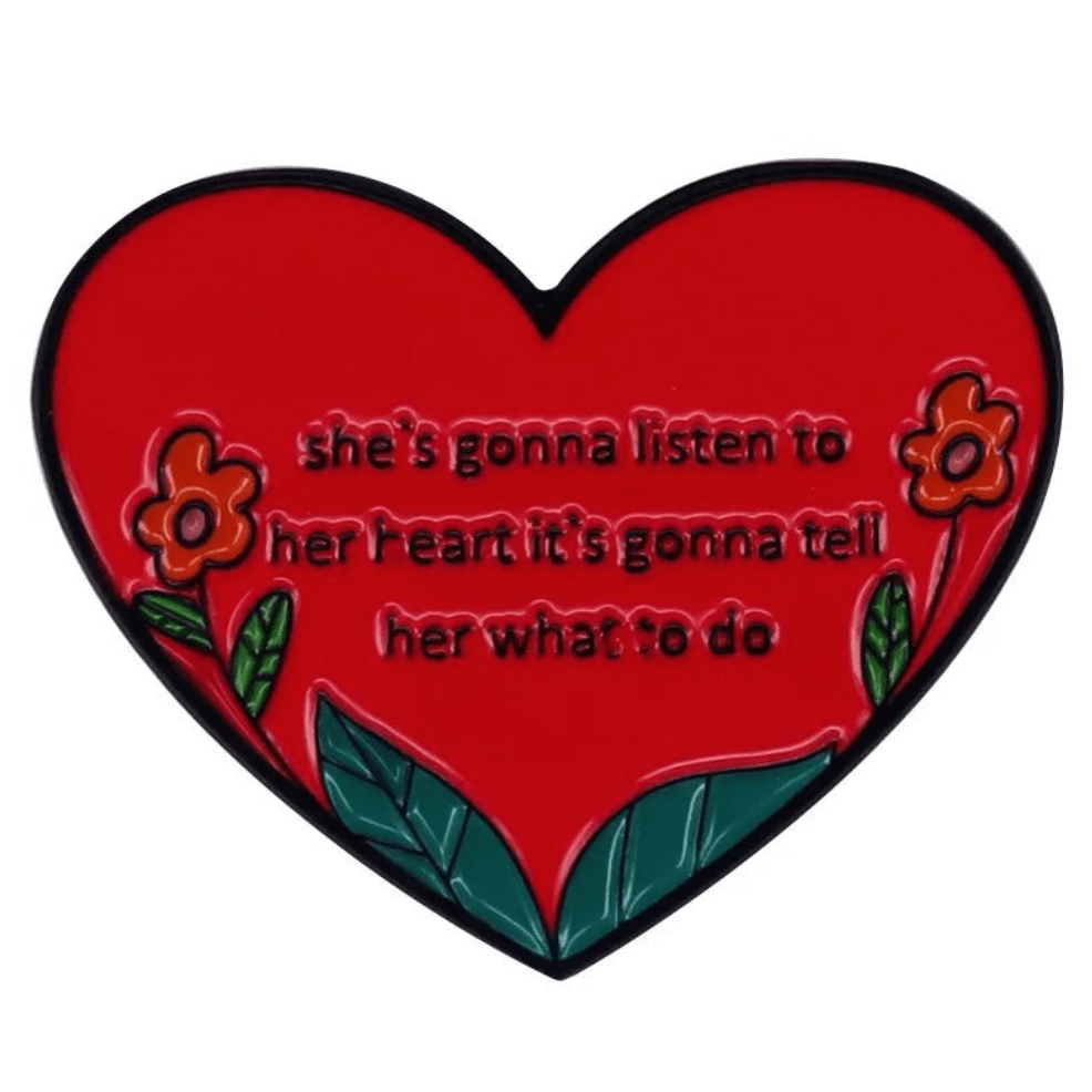 The My Cutesy Era heart enamel pin is red with two orange flowers and green leaves, featuring the text: “she’s gonna listen to her heart, it’s gonna tell her what to do.”.