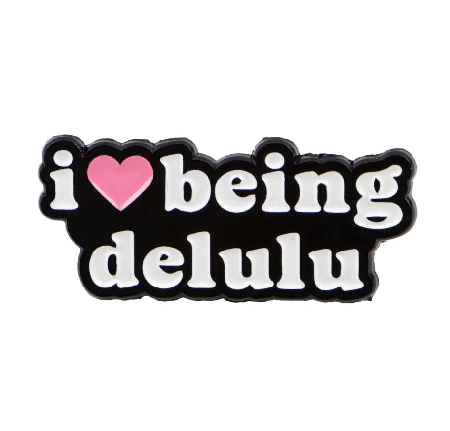 The My Cutesy Era love heart pin displays “i [pink heart symbol] being delulu” in bold white on black—an ideal enamel pin for collectors who enjoy playful, quirky designs.