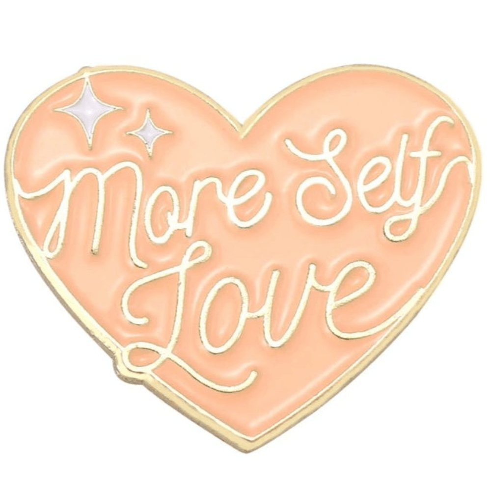 My Cutesy Eras peach heart-shaped enamel pin with gold edges reads More Self Love in handwritten text, accented by two white sparkles—a stylish body positive accessory for daily inspiration.