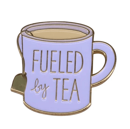 A light purple enamel tea cup pin from My Cutesy Era, shaped like a mug with a teabag charm and FUELED by TEA on the front—a charming accessory for any floral tea cup lover.