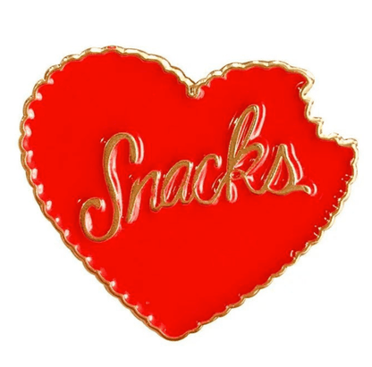A bright red, heart-shaped Snacks pin by My Cutesy Era with a gold outline and gold script. A bite on the upper right gives it a playful touch—perfect for snack lovers!.