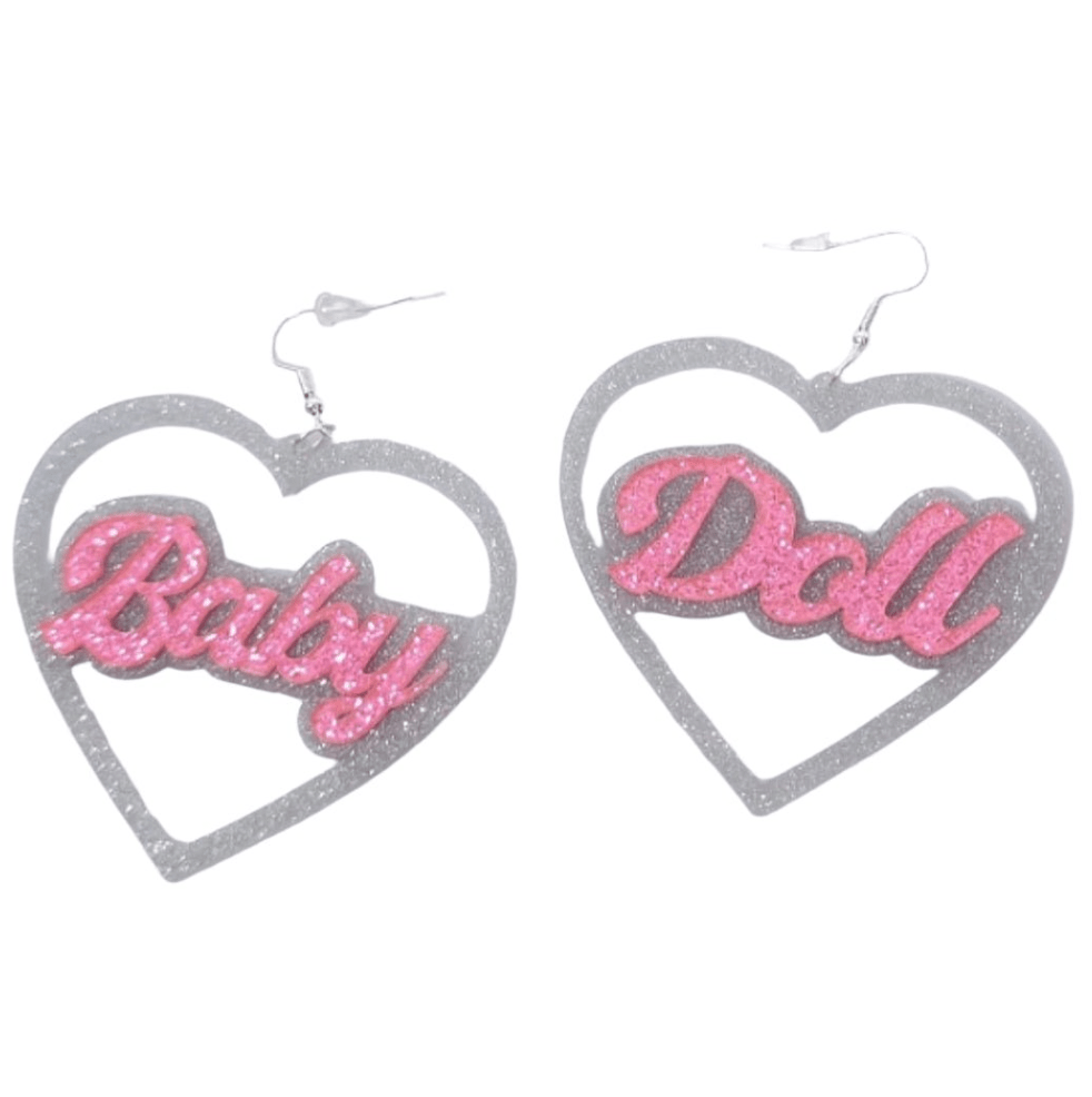A pair of heart-shaped silver and pink glitter earrings from My Cutesy Era, featuring “Baby” and “Doll” in sparkling pink letters—perfect BabyGirl earrings.