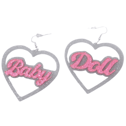 A pair of heart-shaped silver and pink glitter earrings from My Cutesy Era, featuring “Baby” and “Doll” in sparkling pink letters—perfect BabyGirl earrings.