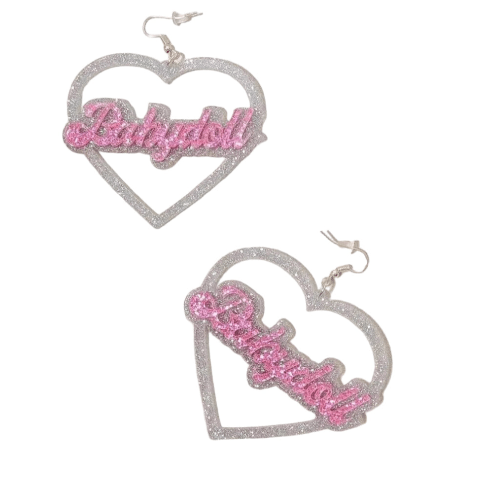 A pair of glittery silver heart-shaped dangly earrings featuring "Babydoll" in pink sparkly letters at the center, by My Cutesy Era, displayed on a white background.