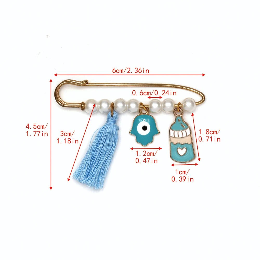 Kiss Me Laters gold safety pin brooch features pearls, a blue tassel, evil eye and hamsa charms, a blue birdcage charm, and red arrows with cm/inch markings. A unique baby shower gift!.