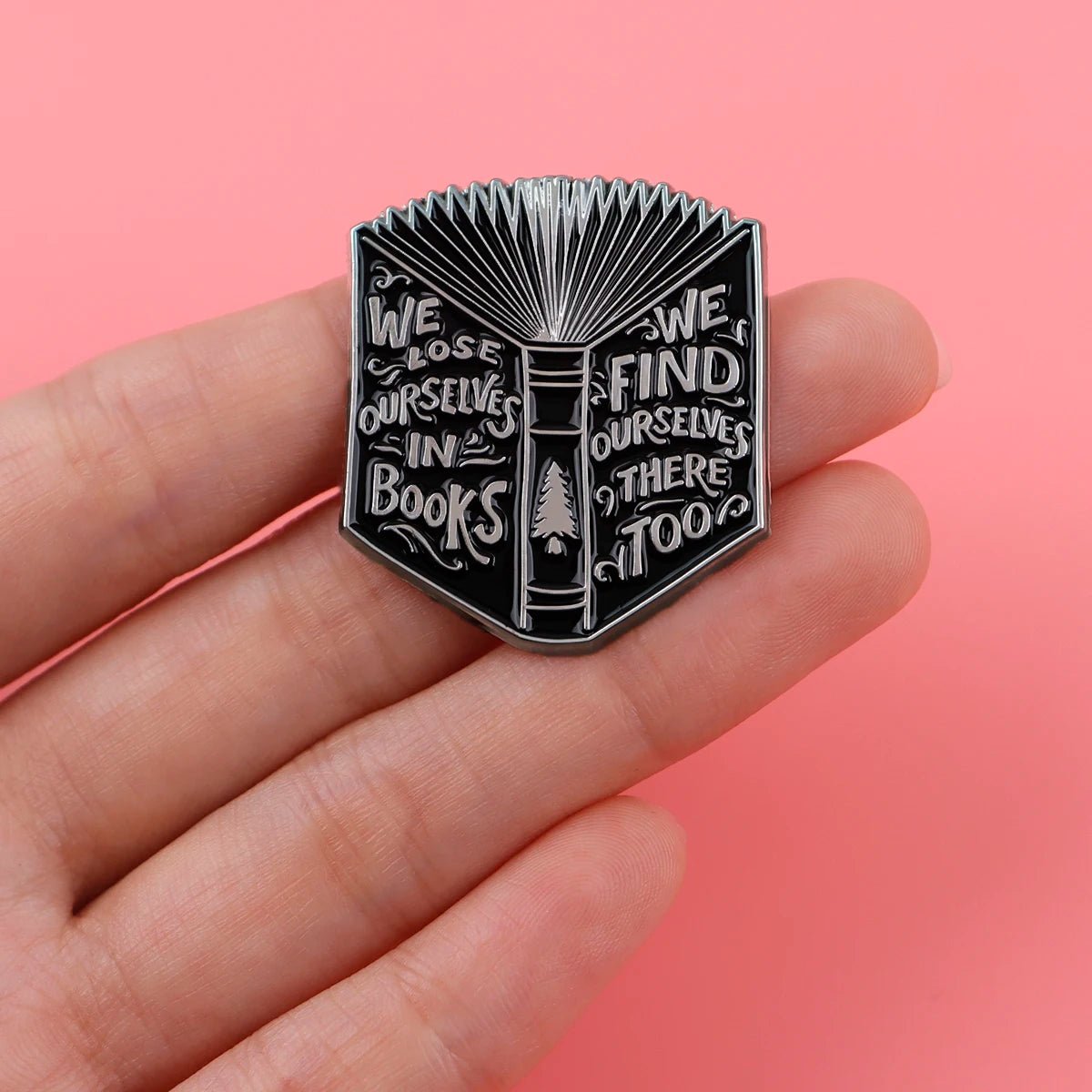 A hand holds an enamel pin shaped like an open book on a pink background. Perfect for Booktroverts, the pin reads: We lose ourselves in books. We find ourselves there too.