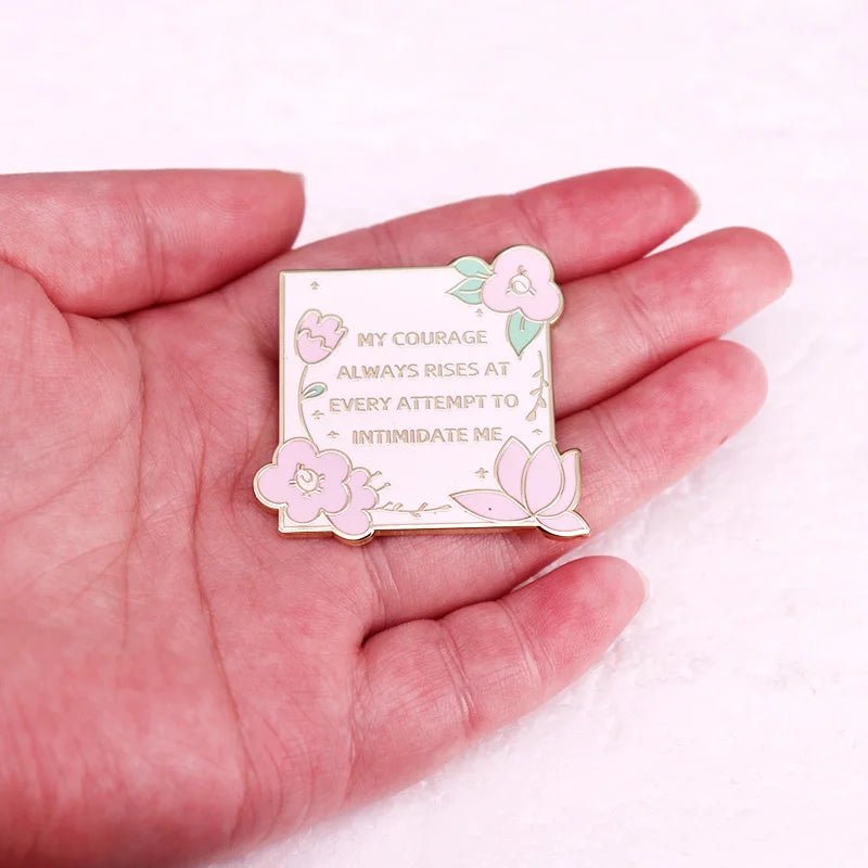 A hand holds a square Kiss Me Later Jane Austen enamel pin with pastel flowers. The gold lettering reads, My courage always rises at every attempt to intimidate me.