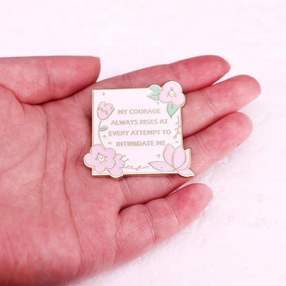 A hand holds a square Kiss Me Later Jane Austen enamel pin with pastel flowers. The gold lettering reads, My courage always rises at every attempt to intimidate me.