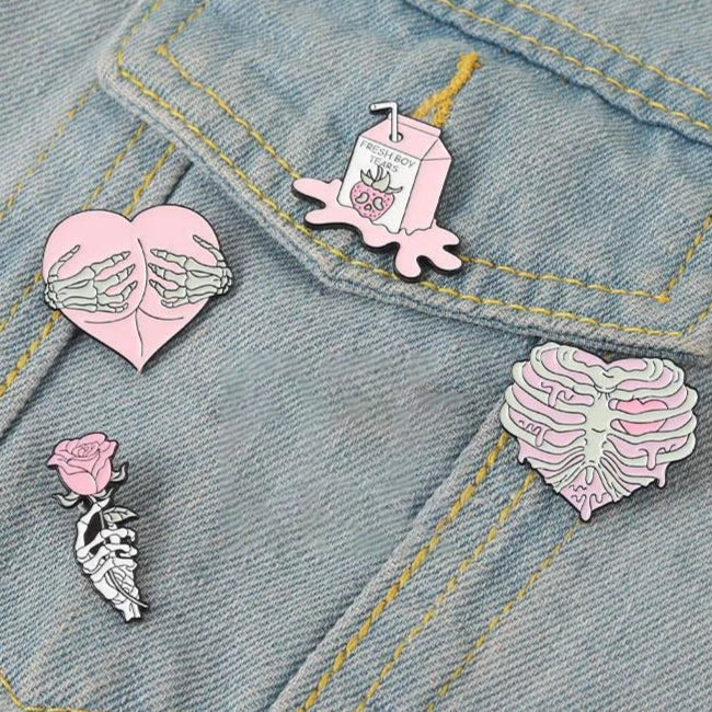 Four enamel pins on light denim: a heart with skeletal hands, a pink milk carton, a skeleton rib cage shaped as a heart, and a skeleton holding a pink rose.