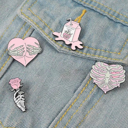 Four enamel pins on light denim: a heart with skeletal hands, a pink milk carton, a skeleton rib cage shaped as a heart, and a skeleton holding a pink rose.