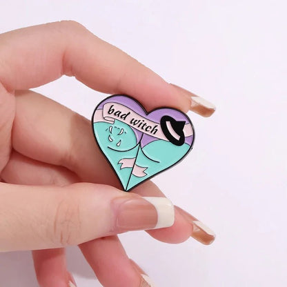 Enamel pin shaped like a heart, featuring bad witch, a witch hat, and a green-skinned character in a pink scarf on a purple background.