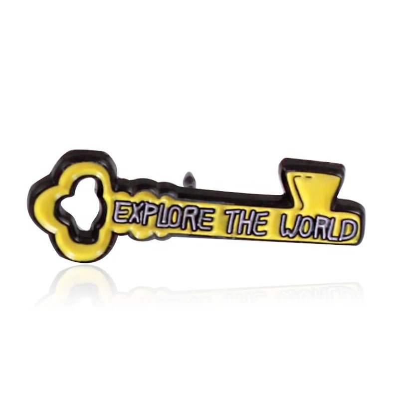A yellow enamel pin shaped like a key, displays EXPLORE THE WORLD in bold capitals on a clean white background.