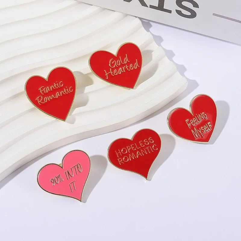 Five heart-shaped red and gold enamel pins featuring phrases like Frantic Romantic, Gold Hearted, Feeling Myself, Hopeless Romantic, and 90% INTO IT, are arranged on a white textured surface.