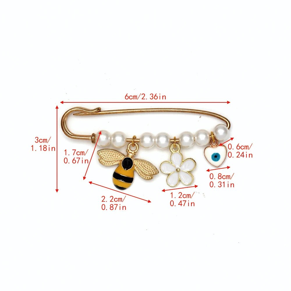The Kiss Me Later gold evil eye pin features white pearls, beads, and charming brooch details—a bee, a white flower, and a blue evil eye—making it a unique baby shower gift. Red lines and numbers display its size in cm and inches.