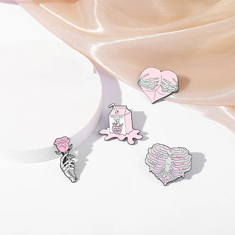 Four pastel enamel pins - a skeleton holding a pink rose, a pink milk carton with dripping liquid, a heart with skeleton hands, and a heart-shaped rib cage. Silk fabric drapes in the background.