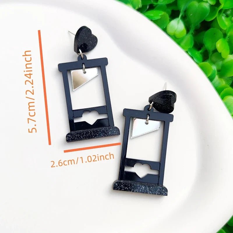 A pair of black guillotine acrylic earrings with cut-out hearts and silver triangle accents from Gay Skate, measuring 5.7 x 2.6 cm, displayed on a white plate beside green leaves.
