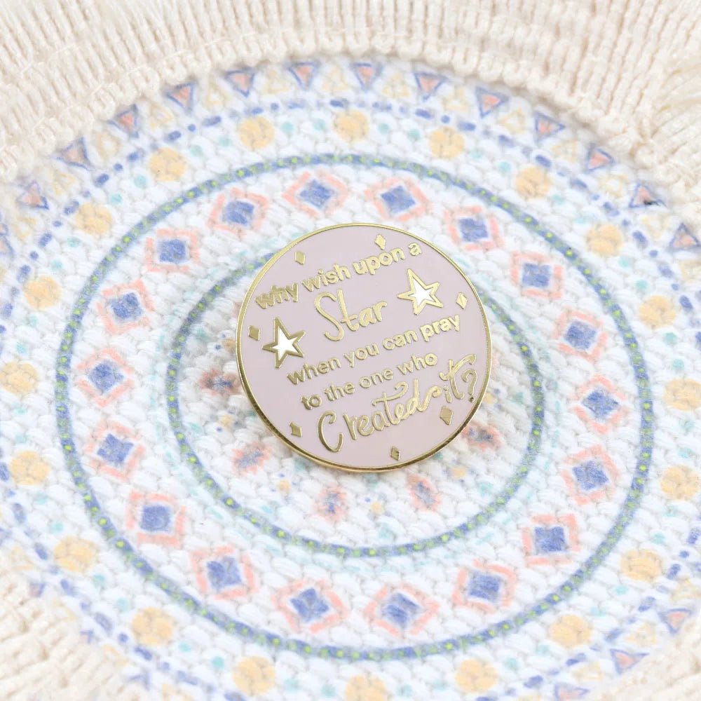 A 3cm x 3cm pink and gold enamel badge reads, Why wish upon a star when you can pray to the one who created it? The round pin is displayed on a textured, colorful patterned fabric.