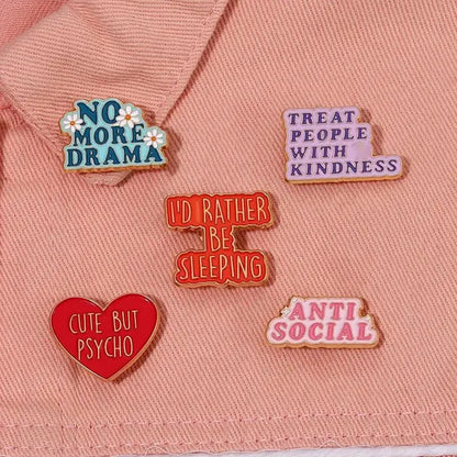 Show off your unique personality with a set of five colorful enamel pins, including a red heart reading Cute But Psycho and other phrases like NO MORE DRAMA and ANTI SOCIAL, all displayed on a pink fabric background.