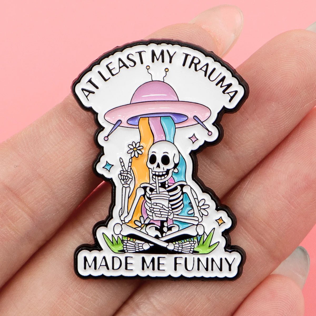 A hand holds an enamel pin featuring a smiling skeleton sitting cross-legged with a rainbow and UFO. The pin says “At least my trauma made me funny” on pink—a quirky alien-themed addition to your collection.
