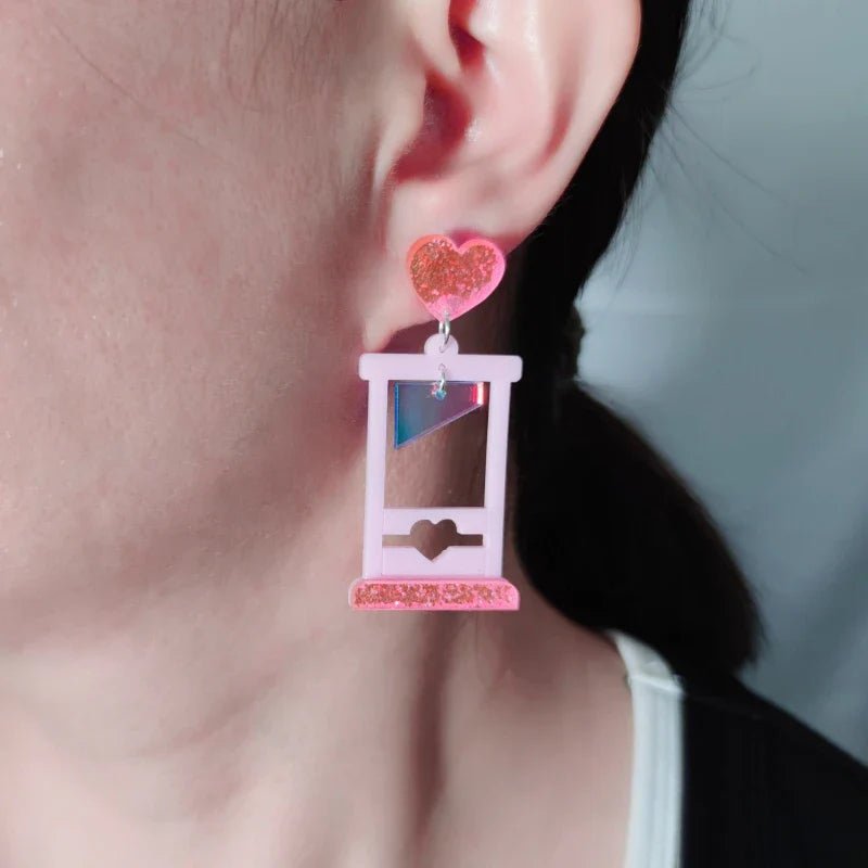 A close-up of a persons ear wearing Gay Skates guillotine acrylic earrings, shaped like a small pink guillotine with a glittery pink heart at the top, against a softly blurred background.