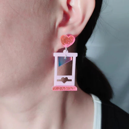 A close-up of a persons ear wearing Gay Skates guillotine acrylic earrings, shaped like a small pink guillotine with a glittery pink heart at the top, against a softly blurred background.