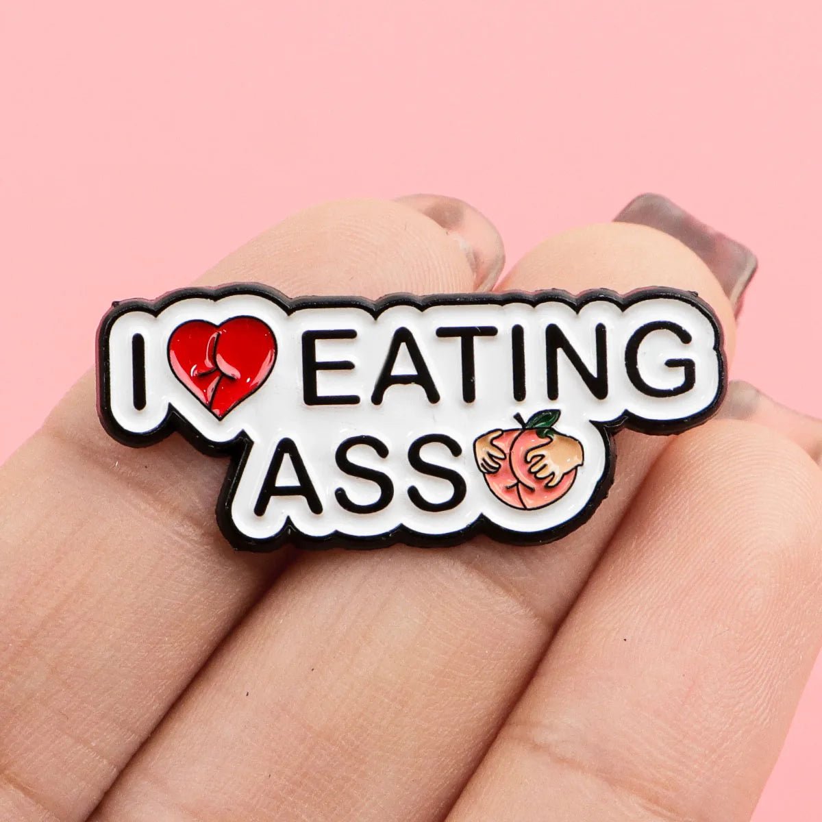 A hand holds a Kiss Me Later peach pin that says I Love Eating Ass with a red heart and cartoon peach, 3.5cm x 1.5cm, against a pink background.