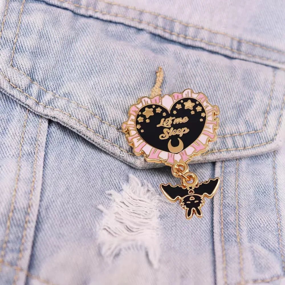 Enamel pin features a pink and black heart reading “Let Me Sleep,” with stars, a crescent moon, and a tiny bat charm—adding a dreamy vibe when pinned to a ripped light denim jacket.