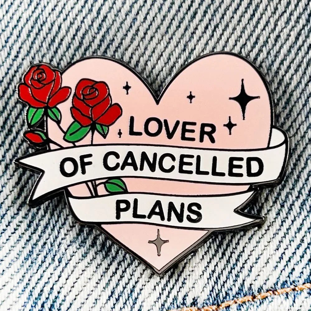 Kiss Me Later’s heart-shaped enamel pin, featuring red roses and a Lover of Cancelled Plans banner, sits on blue denim. Black stars highlight the pink background, making this pin a delightful statement piece.