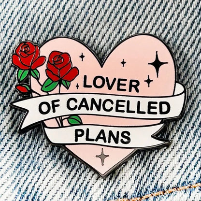 Kiss Me Later’s heart-shaped enamel pin, featuring red roses and a Lover of Cancelled Plans banner, sits on blue denim. Black stars highlight the pink background, making this pin a delightful statement piece.