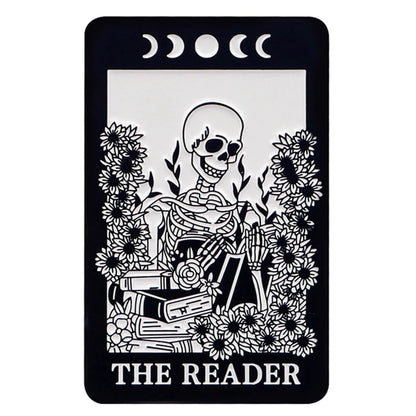 Kiss Me Later’s tarot card-style illustration, “The Reader,” features a smiling skeleton amid blooming flowers and stacked books, with spooky vibes from three crescent moons and a full moon above—all in striking black and white.