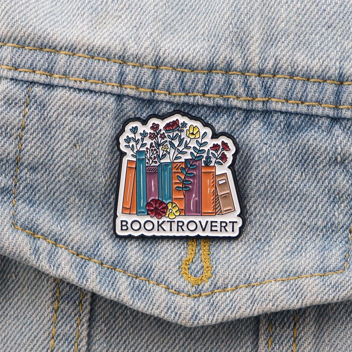 Colorful enamel pin for denim shows books with blooming flowers and BOOKTROVERT below—a perfect accessory for fans of fictional characters and public libraries.