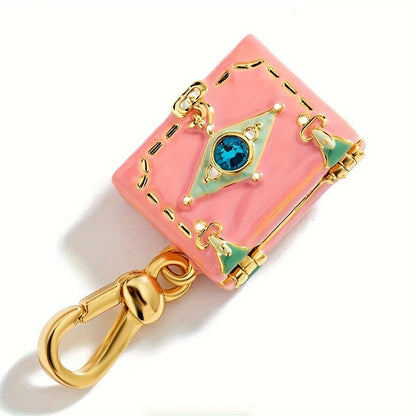 A pink, suitcase-shaped gold enamel pendant by features green accents, a blue center gem, and a gold clasp for attaching to a bracelet or necklace, photographed on a white background.
