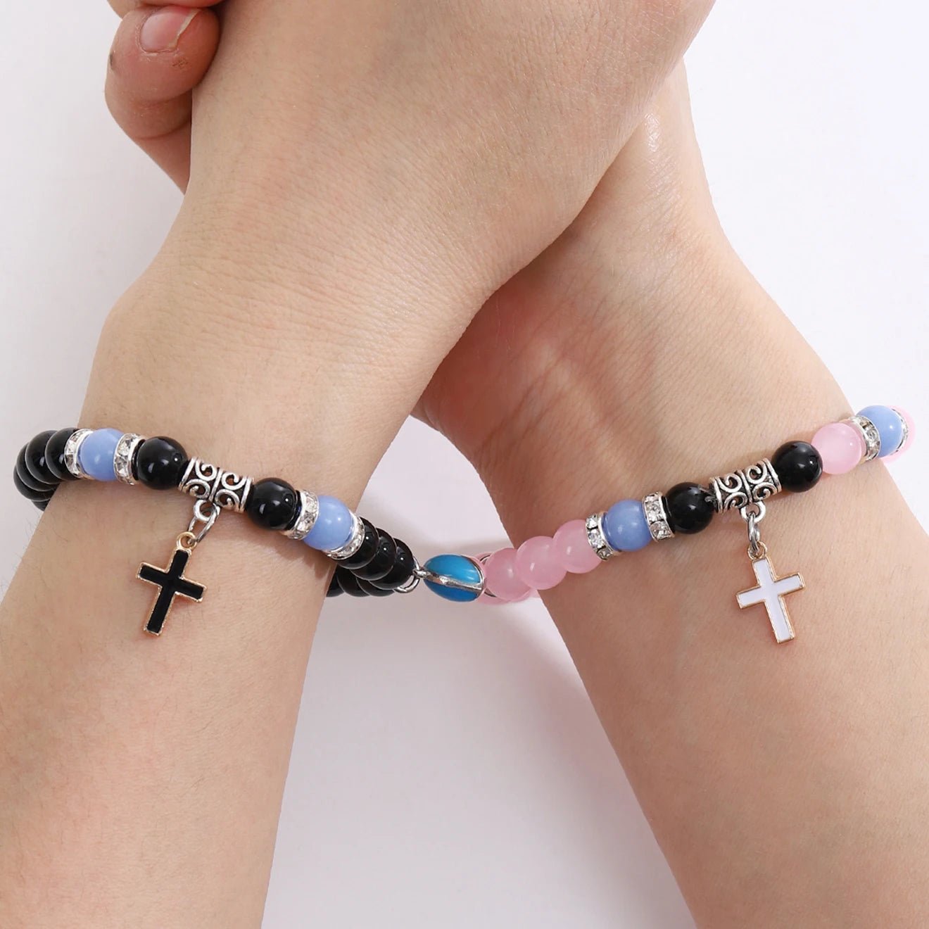 Two hands crossed at the wrist, each wearing a Kiss Me Later beaded bracelet with silver accents and small cross charms—one bracelet features a glow-in-the-dark cross pendant, making it a meaningful baptism gift.