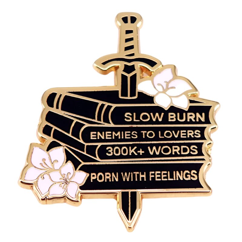 enamel pin features a gold and black book stack pierced by a sword, with book spines labeled: Slow Burn, Enemies to Lovers, 300K+ Words, and With Feelings. Pink flowers accent the sides.