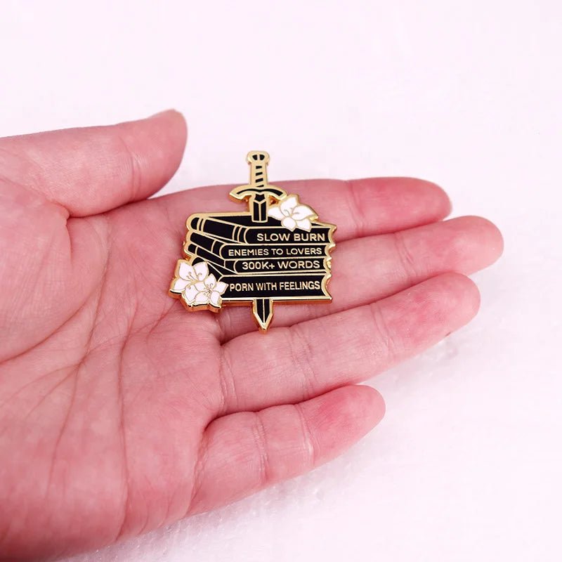 A hand holds an enamel pin: a dagger through stacked books with white flowers. The spines read Slow Burn, Enemies To Lovers, 300K+ Words, and With Feelings.