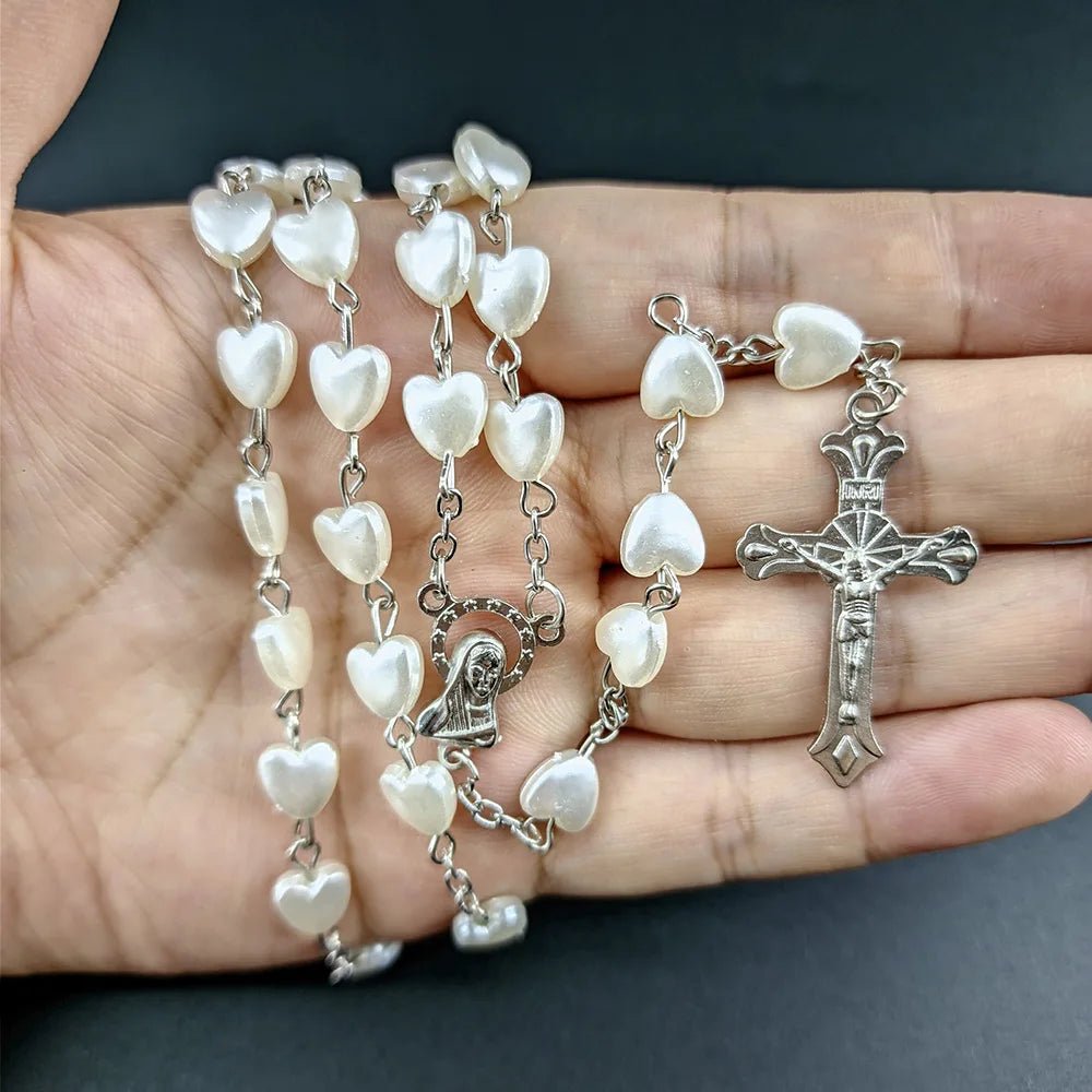 A hand holds a My Cutesy Era rosary necklace with white, heart-shaped beads, a small Virgin Mary medallion, and a metallic crucifix against a dark background.