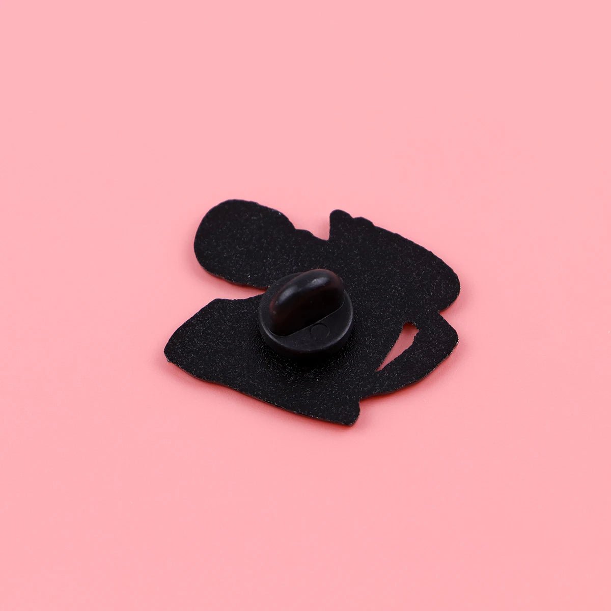 A small black bean sits on an abstract felt cutout next to a quirky Funny Skeleton enamel pin, all arranged on a solid pink background.
