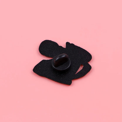 A small black bean sits on an abstract felt cutout next to a quirky Funny Skeleton enamel pin, all arranged on a solid pink background.