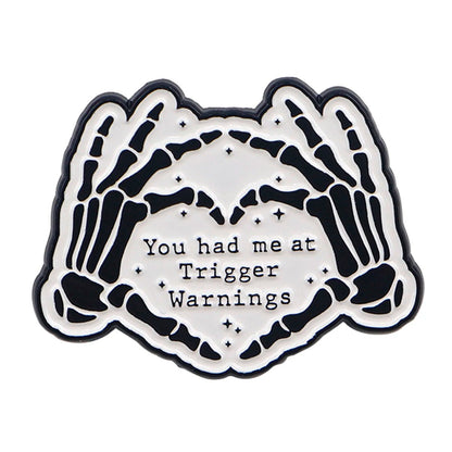 The Kiss Me Later enamel pin features two skeleton hands forming a heart around the words You had me at Trigger Warnings on a white background—perfect for adding spooky vibes to your style.