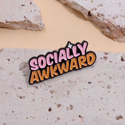 A pink and orange enamel pin  featuring the words SOCIALLY AWKWARD, rests on a light stone-textured surface with additional stone pieces in the background.