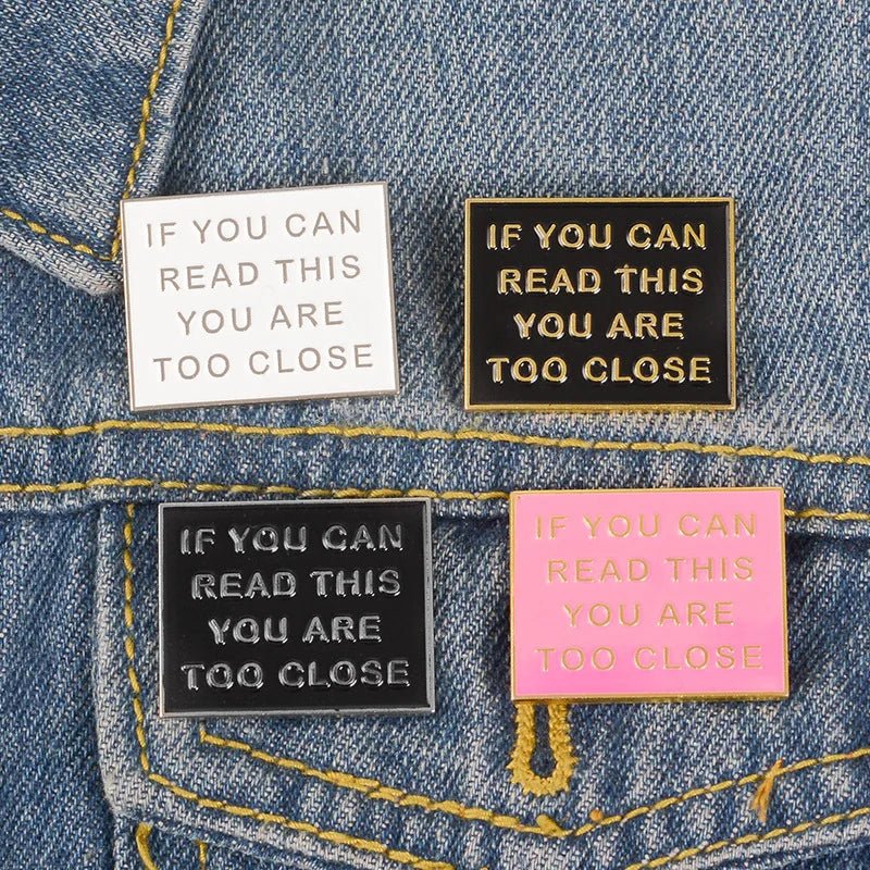 Add quirky flair with this enamel square pins—set of four on blue denim, each stating “IF YOU CAN READ THIS YOU ARE TOO CLOSE.” Available in white, black, gold, and pink with bold text for a playful warning.