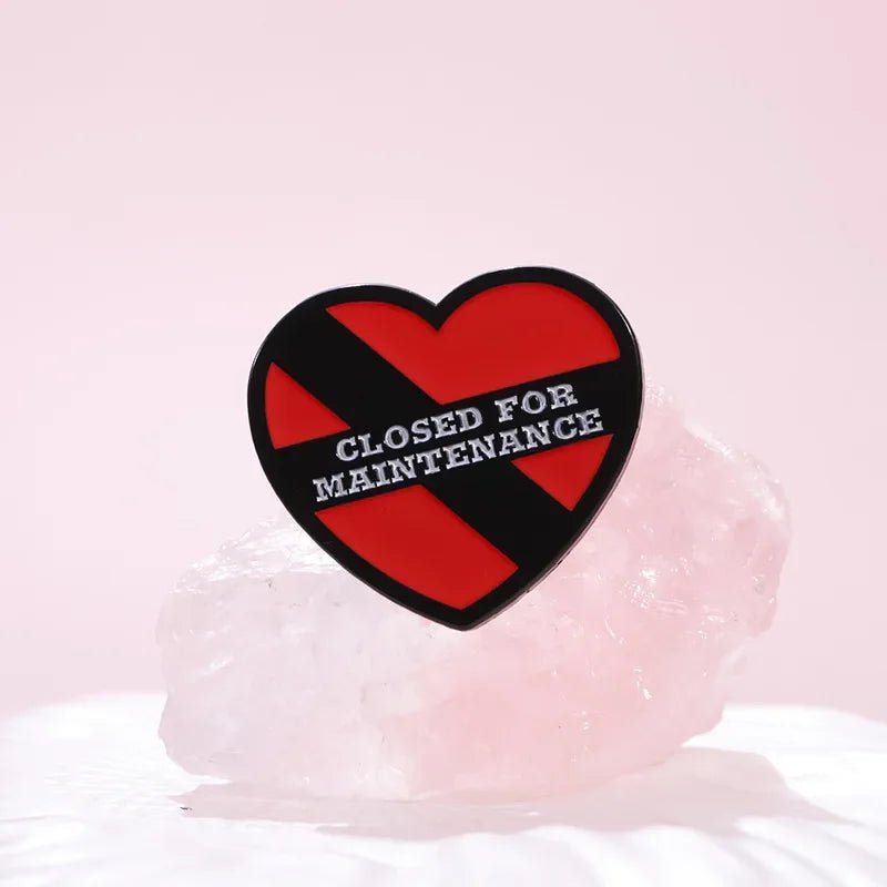 Red heart enamel pin features a black diagonal stripe and “CLOSED FOR MAINTENANCE” in white, displayed on a translucent pink crystal with a soft pink background.