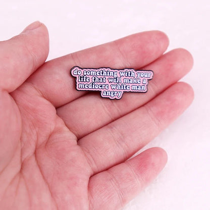 A hand displays a My Cutesy Era feminist enamel pin reading do something with your life that will make a mediocre white man angry against a soft pink background.