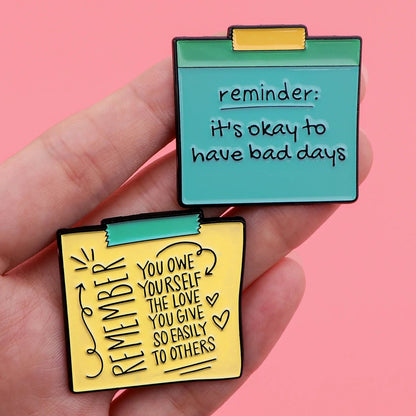 A hand holds two colorful pins shaped like sticky notes, each with positive affirmations: one reads, reminder: it’s okay to have bad days, and the other promotes self empowerment on a gentle pink background.