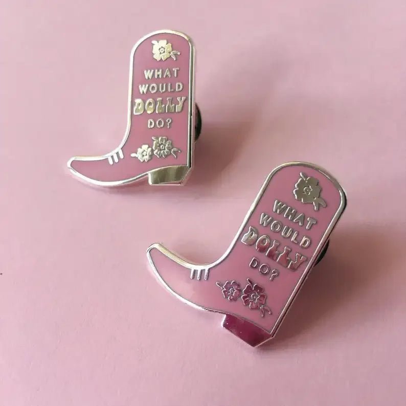 Two pink and gold cowboy boot enamel pins outlined in silver with What Would Dolly Do? and floral details, displayed on a pastel pink background—perfect for any Dolly fan’s enamel pin collection.